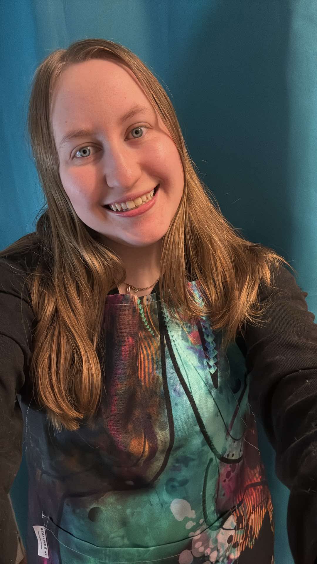Blonde woman smiling at the camera while wearing the colorful Neuro-Spicy Brain apron, featuring abstract artistic designs in teal, pink, and orange.