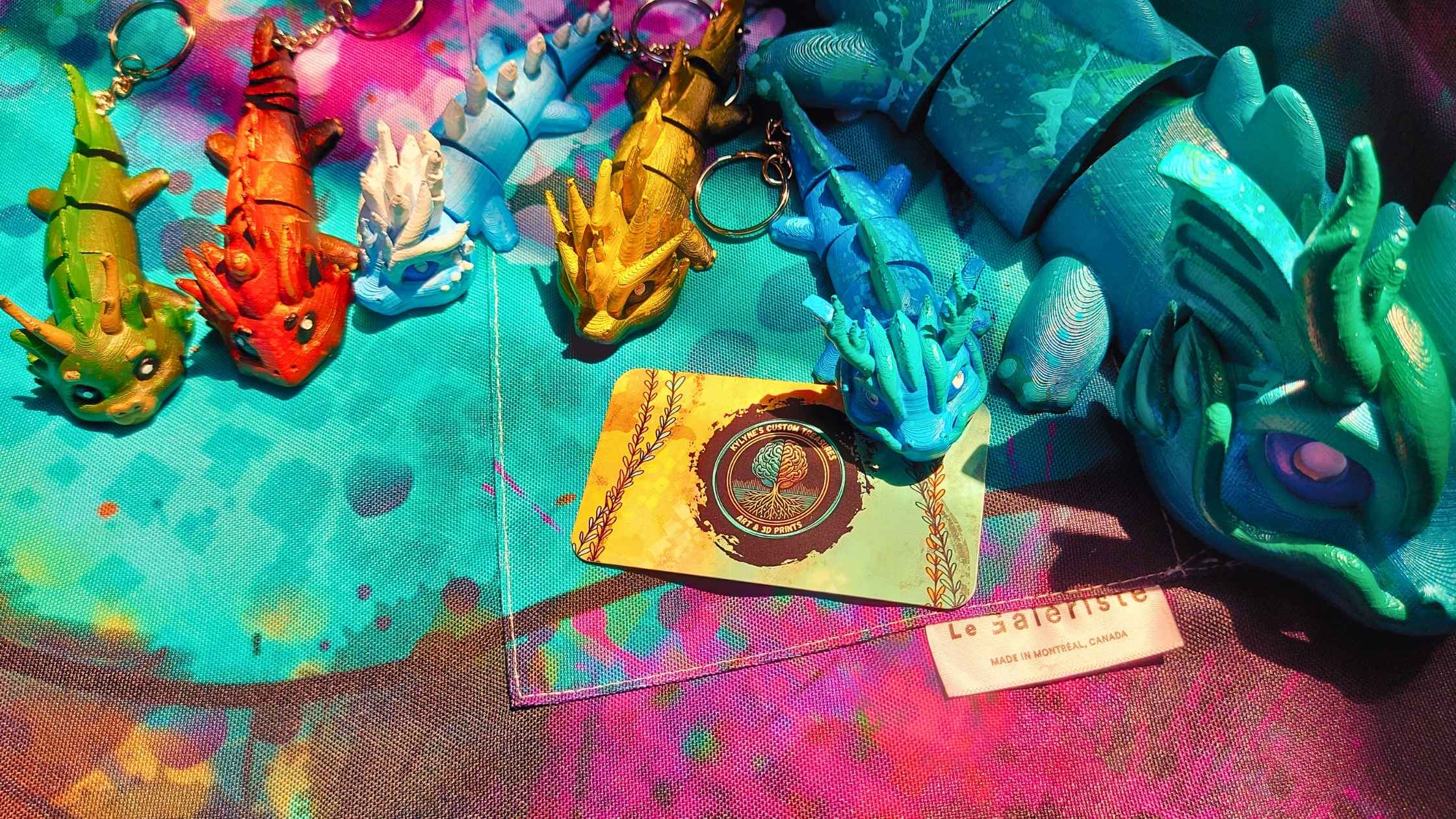 Hand-painted dragon keychains in a variety of colors arranged on a vibrant, splatter-painted fabric background, next to a Kylyne’s Custom Treasures business card and a Le Galeriste label. Custom art by Carolyne D. Landry.