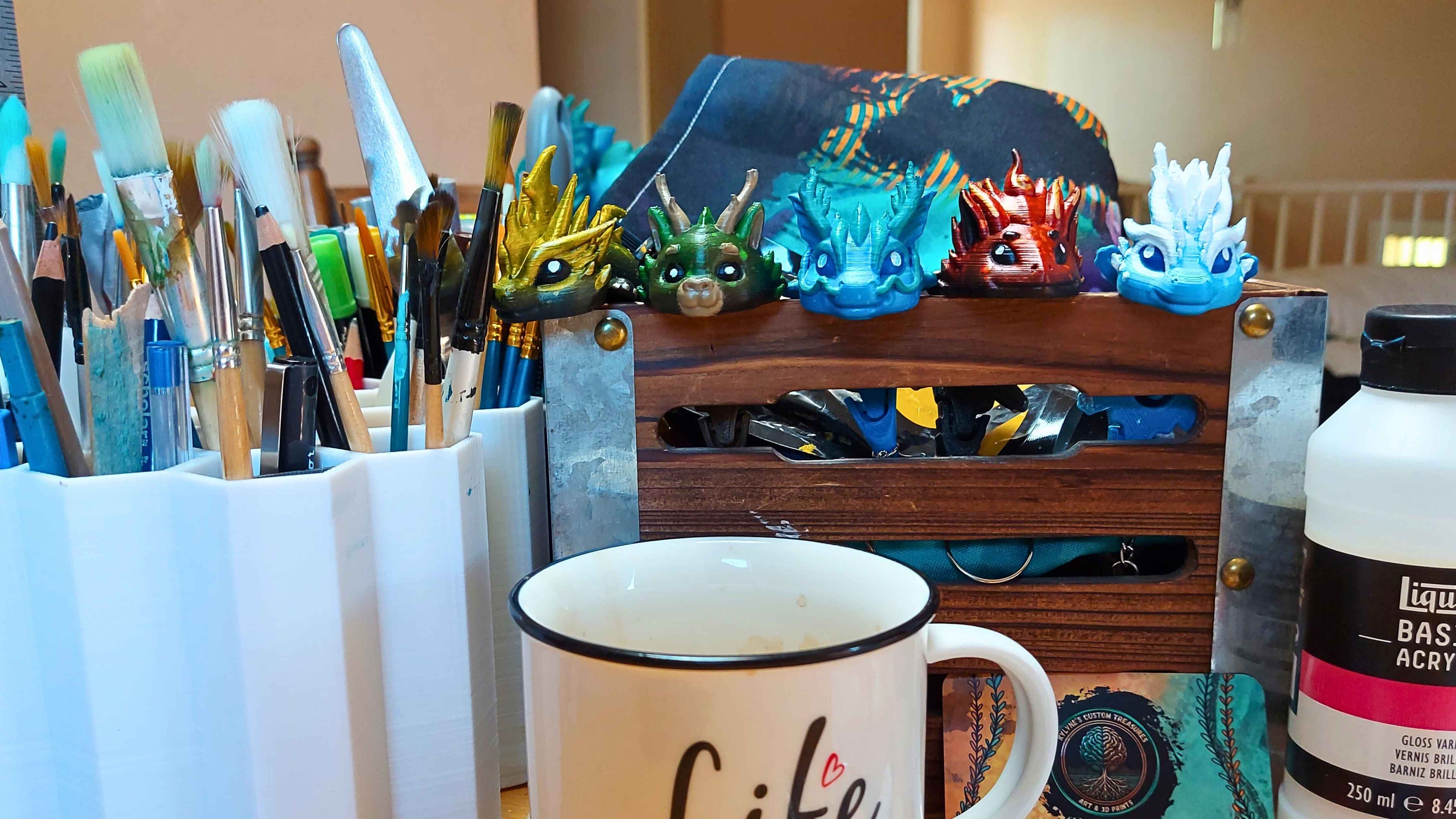 Artist’s workspace featuring a white mug with the word ‘Life’, paintbrushes, and a row of colorful hand-painted dragon keychain heads by Kylyne’s Custom Treasures. The dragons are displayed on a wooden organizer, with art supplies and a vibrant cloth in the background.
