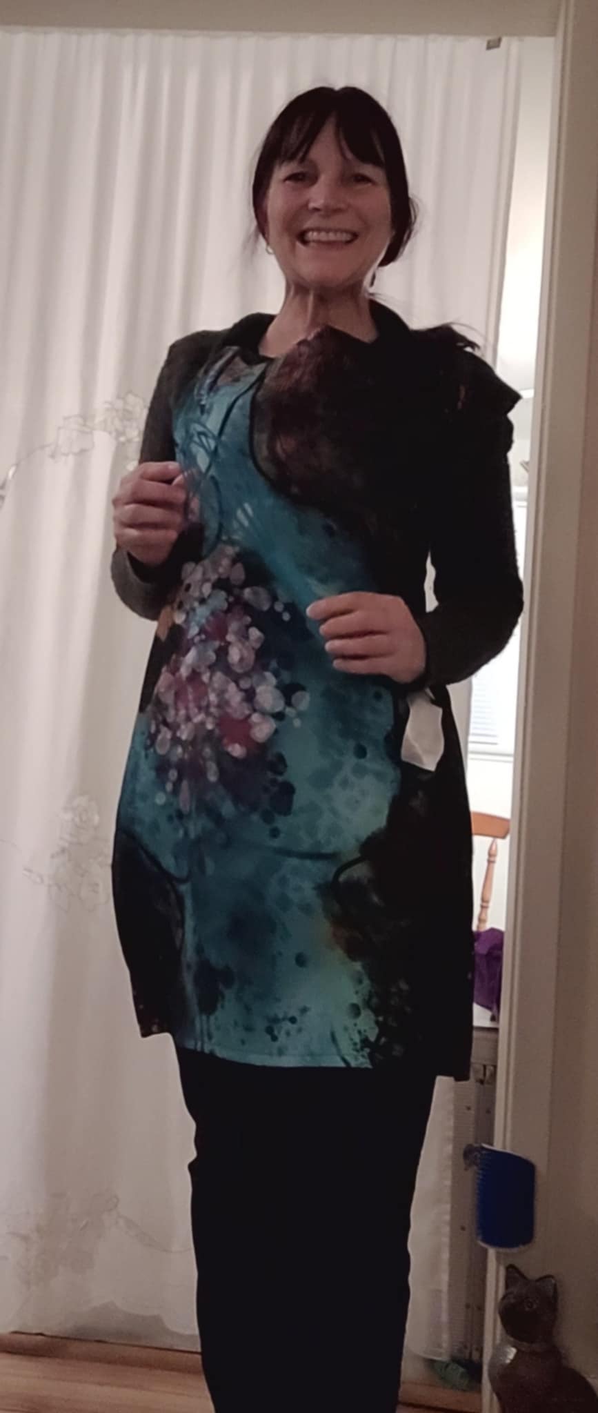 A smiling woman wearing the Neuro-Spicy Brain apron, showcasing its vibrant teal and purple abstract design in a home setting.