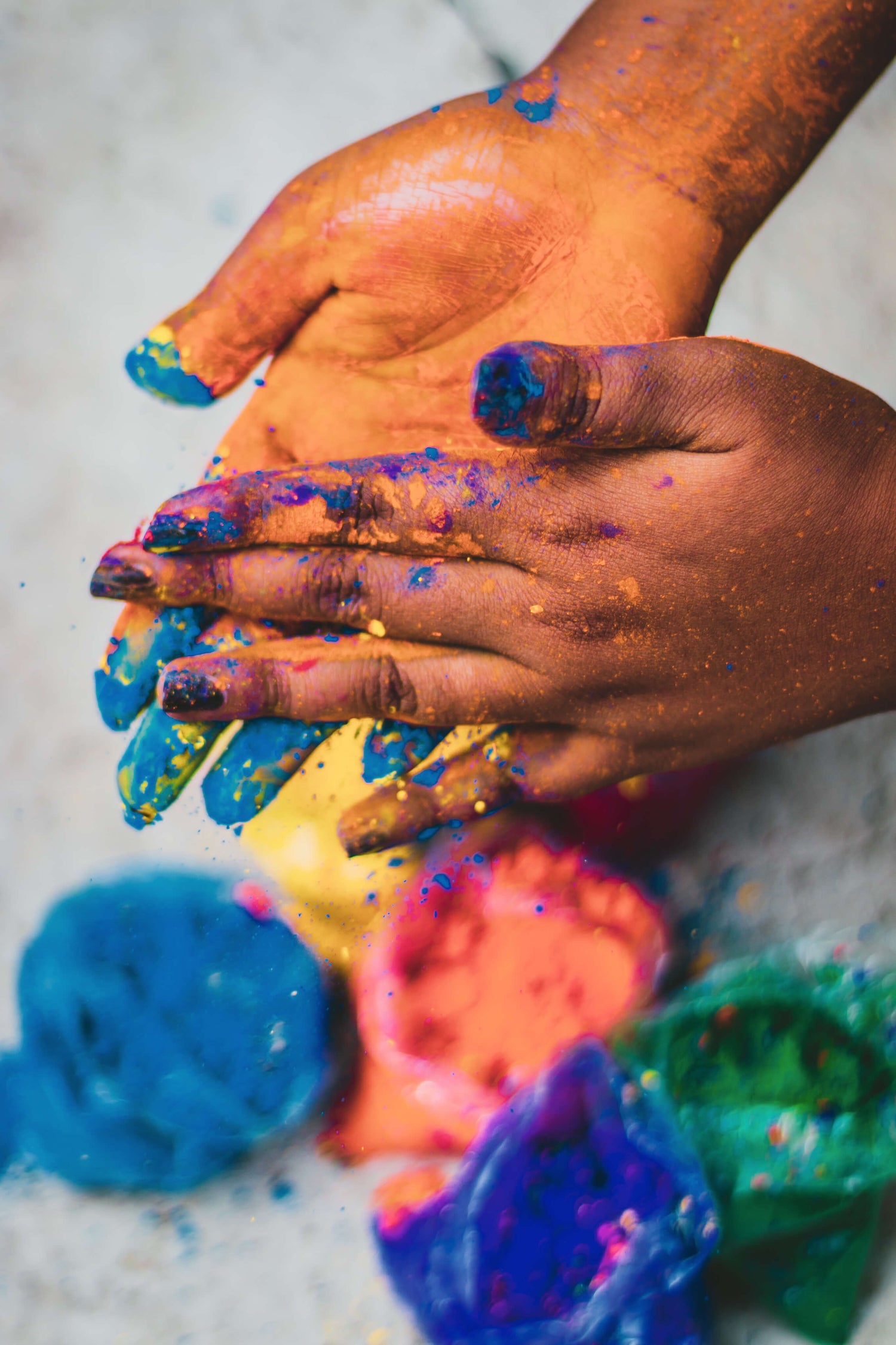 Hands covered in colorful paint, with open paint containers in blue, green, orange, and purple visible below, capturing the creative and messy process of making art.