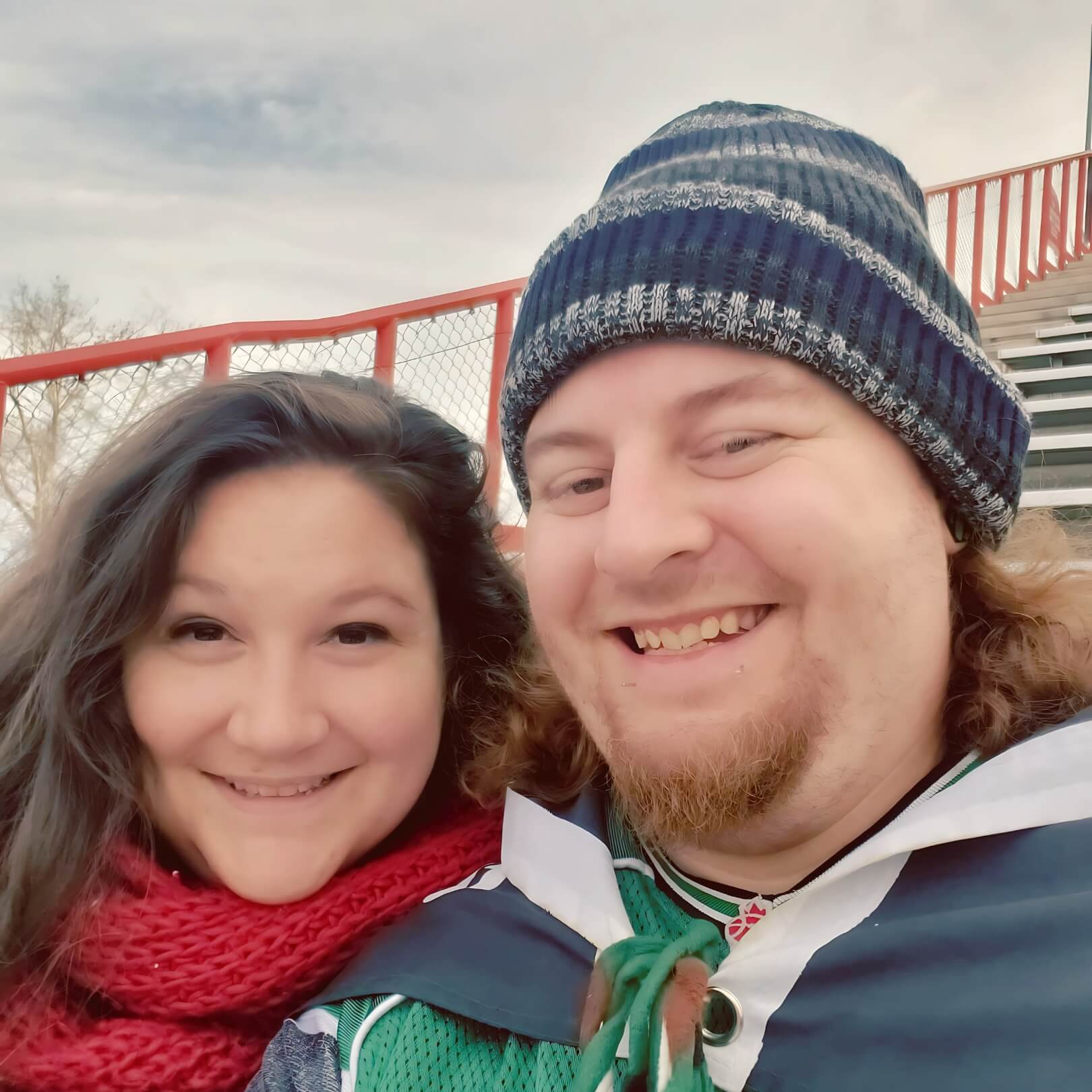 Carolyne and Kyle smiling together at a Riders football game, bundled up in winter clothing with red stadium railings in the background.