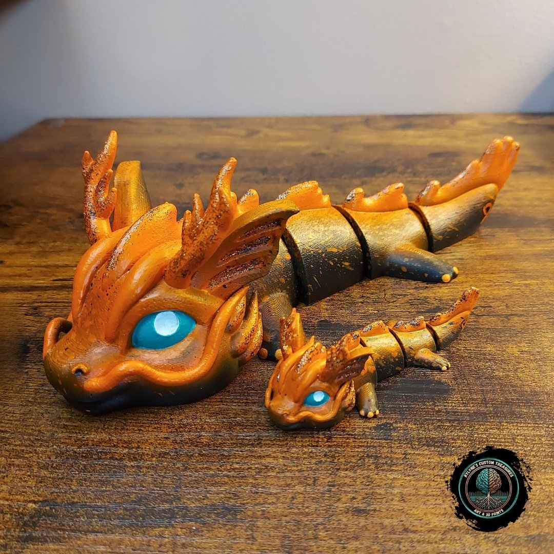 Custom hand-painted articulated dragon figurines in orange and black with blue eyes, large and keychain size, 3D-printed by Kylyne’s Custom Treasures, displayed on a wood background.