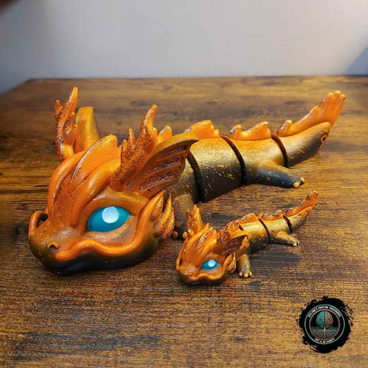 Custom hand-painted articulated dragon figurines in orange and black with blue eyes, large and keychain size, 3D-printed by Kylyne’s Custom Treasures, displayed on a wood background.