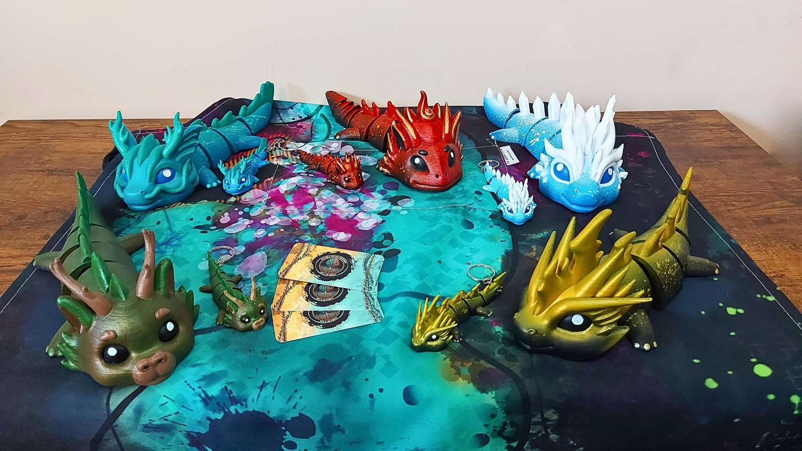 Group display of hand-painted, 3D-printed articulated elemental dragon figurines by Kylyne’s Custom Treasures, including Vaylen, Eryndra, Kaelix, Dasyra, and Zoryn in various sizes on a colorful game board with business cards. Sensory-friendly art for neurodivergent collectors.