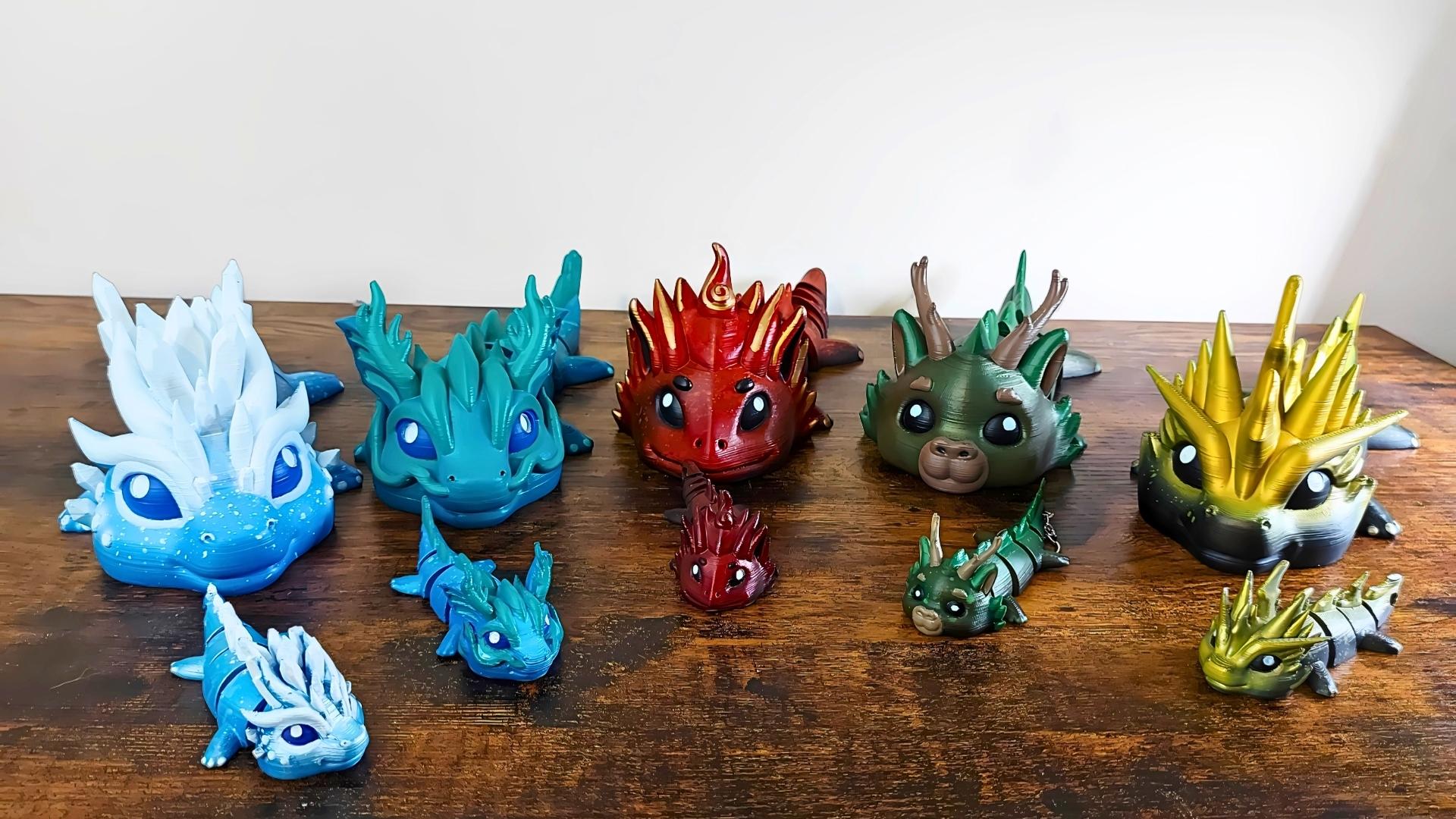 Group of five Elemental Dragons of Drakora—hand-painted and 3D-printed collectible dragon figurines by Kylyne’s Custom Treasures, designed by Biocraftlab. Includes blue, red, green, gold, and white dragons in various sizes, arranged on a wooden table.