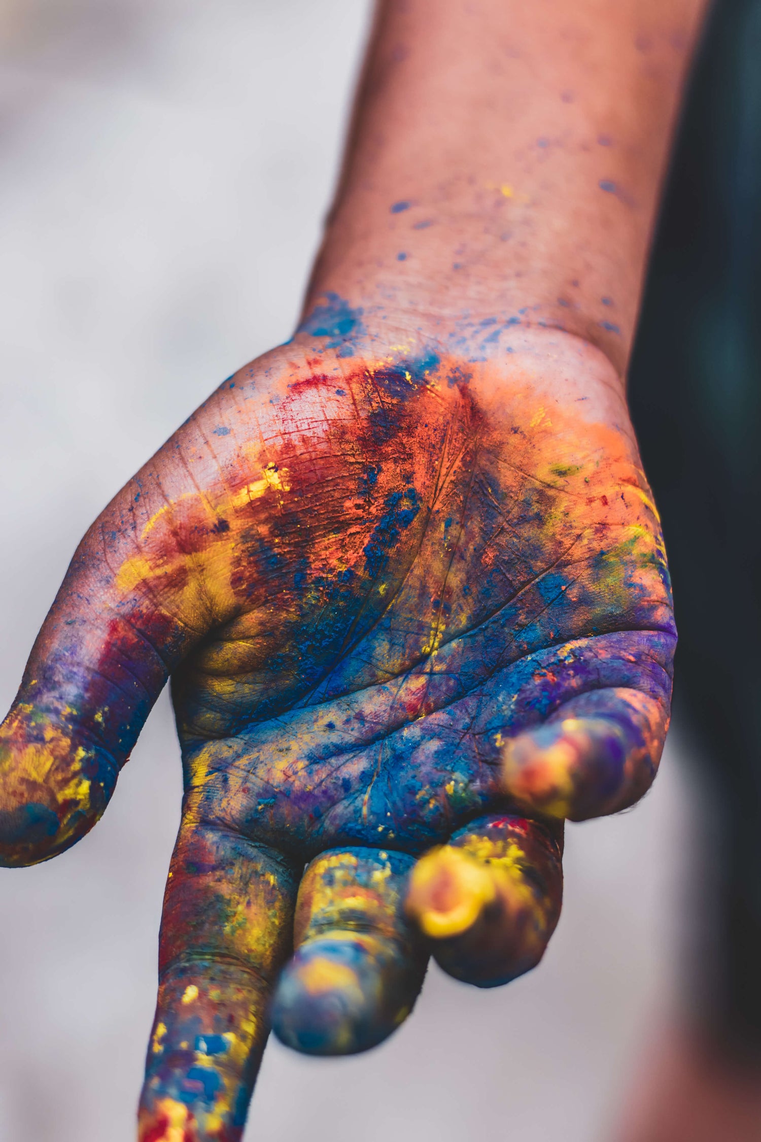 A close-up of an open hand covered in vibrant splashes of blue, orange, yellow, and red paint, capturing the creative and expressive process of making art.