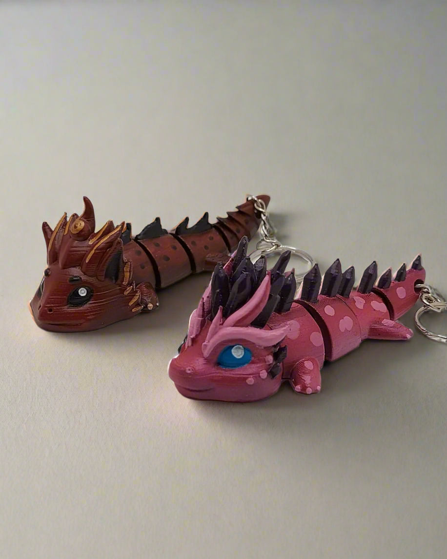 Two custom hand-painted dragon keychains by Kylyne’s Custom Treasures—one brown with gold accents, one pink with purple spikes and blue eyes.