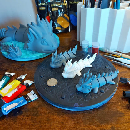 Behind-the-scenes worktable with primed dragon keychains for the Paint Your Dragon Kit, art supplies, and a larger Kaelix dragon in the background.