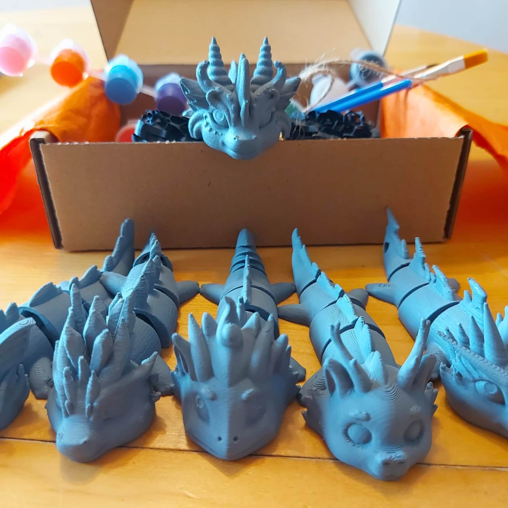 Paint Your Dragon Kit box with paint pods and brushes behind a lineup of six primed dragon keychain examples.