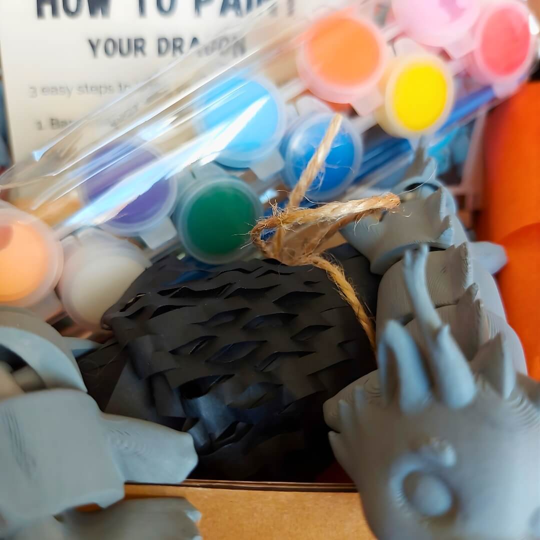 Close-up inside the Paint Your Dragon Kit box showing acrylic paint pods, packing paper, and primed dragon pieces.