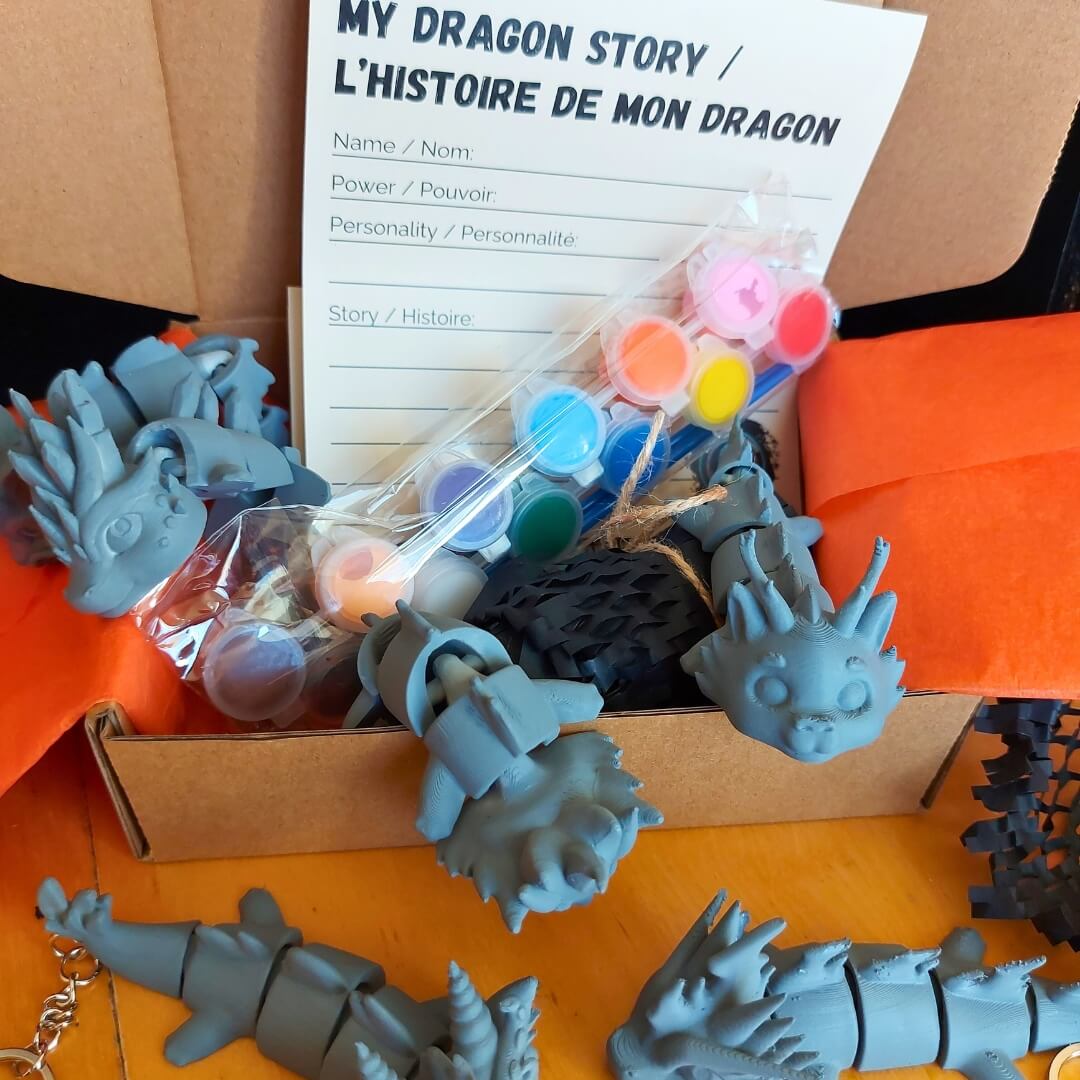Open Paint Your Dragon Kit box showing paint pods, “My Dragon Story” card, packing paper, and multiple primed dragon keychains.