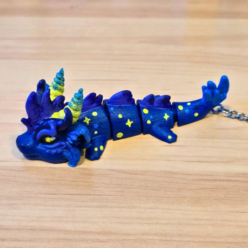 Customer-painted finished example from the Paint Your Dragon Kit: a dark blue dragon keychain with yellow stars on a wooden surface.