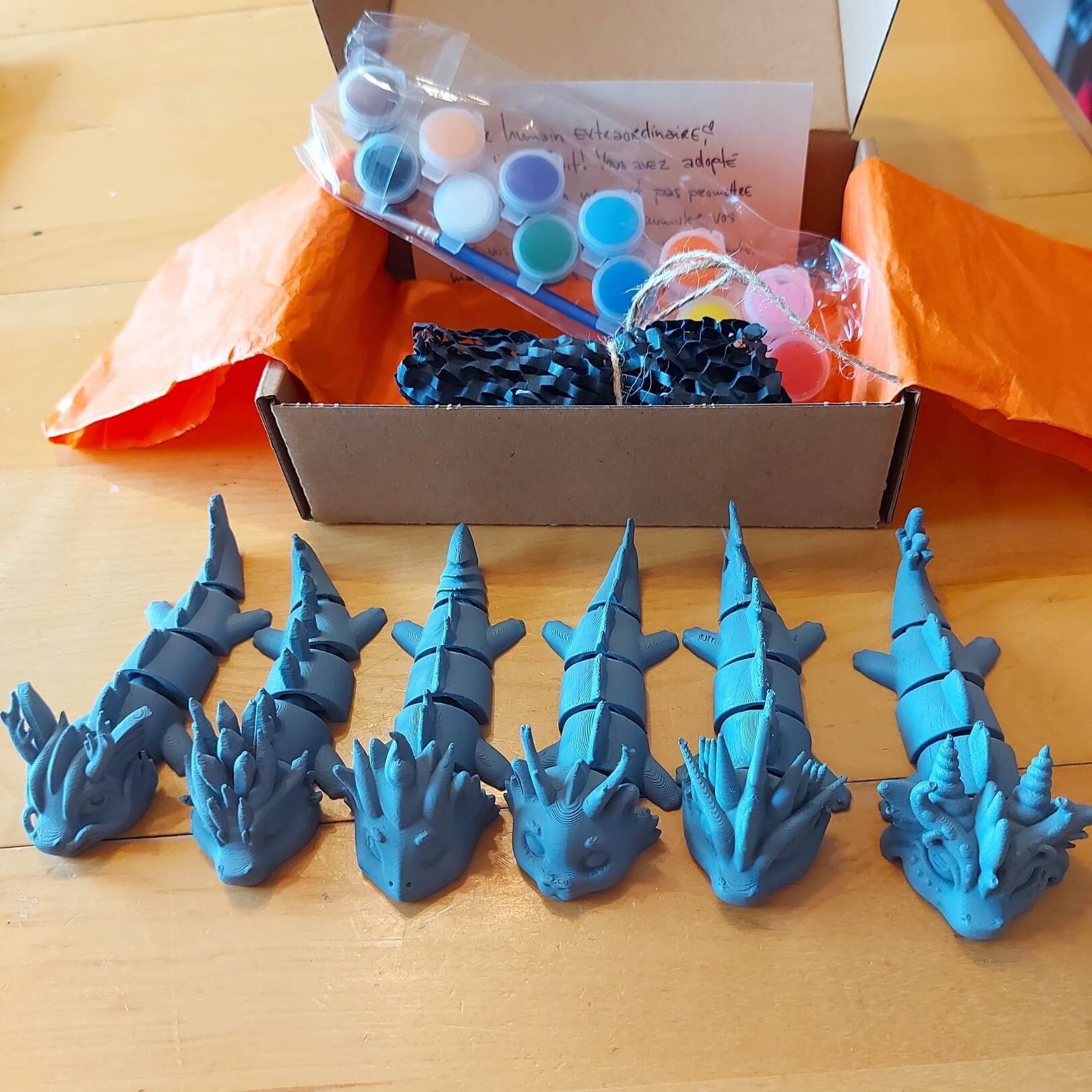 Paint Your Dragon Kit box with paint pods and instructions behind a lineup of six primed dragon keychain examples.