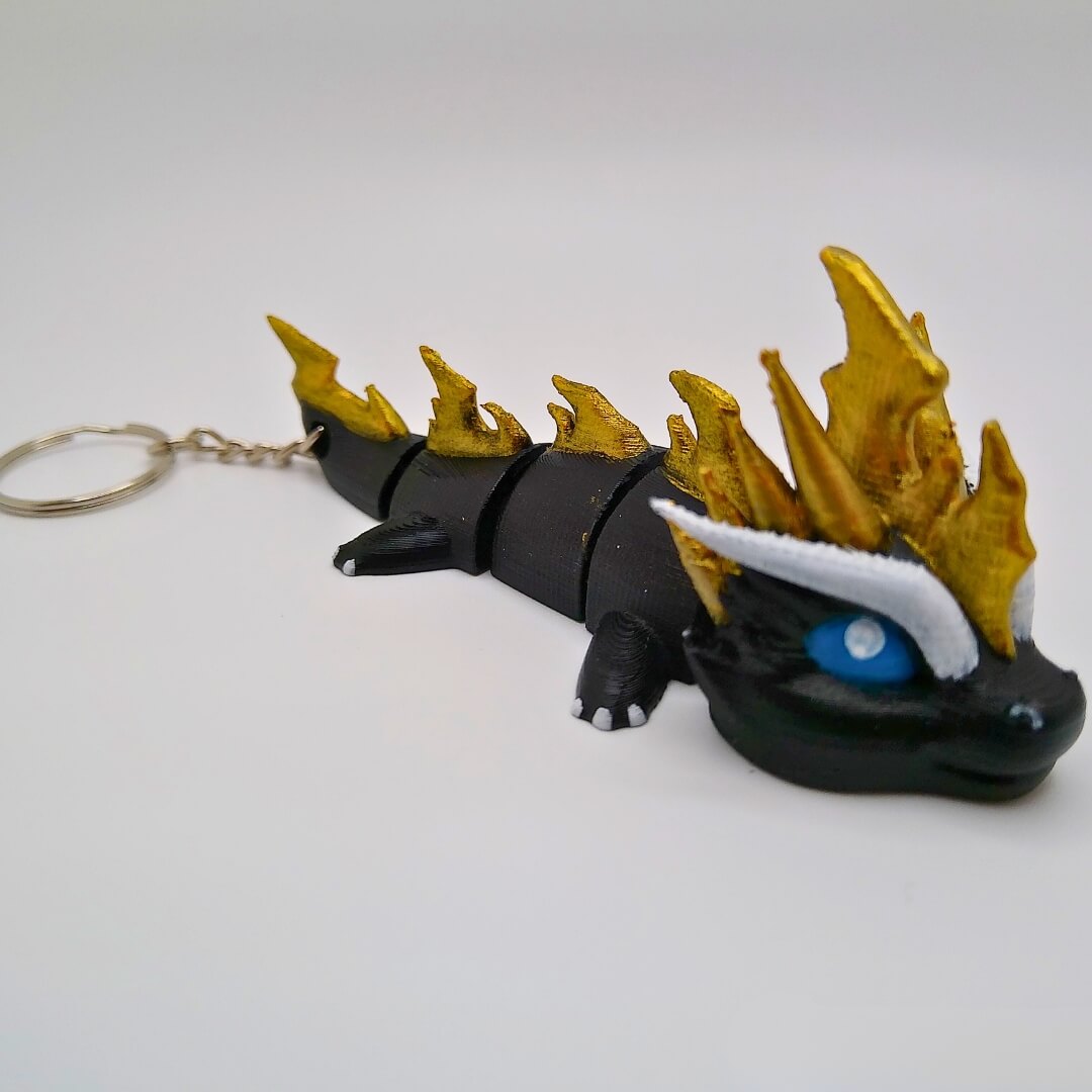 Finished example of a hand-painted dragon keychain from the Paint Your Dragon Kit, black with gold spikes and blue eyes, on a white background.