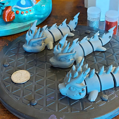 Several primed dragon keychains prepared for the Paint Your Dragon Kit, with a partially painted showgirl dragon in the background.