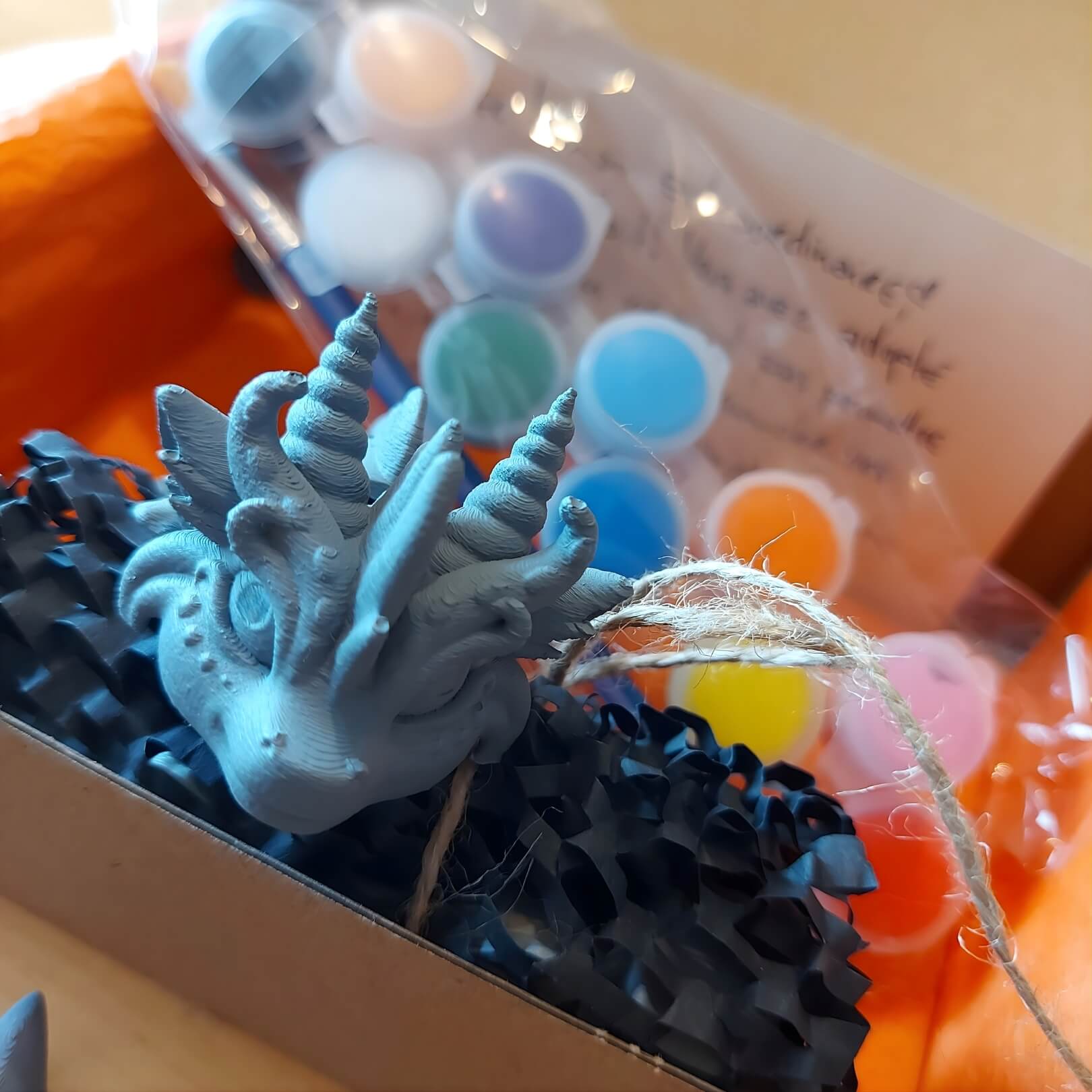 Close-up of Zephir, a primed dragon keychain, inside the Paint Your Dragon Kit box with paint pods and packing paper.